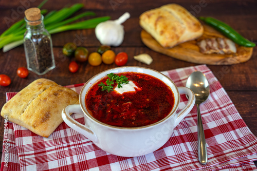 Borscht with sour cream in a white plate on a wooden table. The table also contains bread, bacon, green onions, peppers and garlic. Traditional Ukrainian and Russian cuisine.