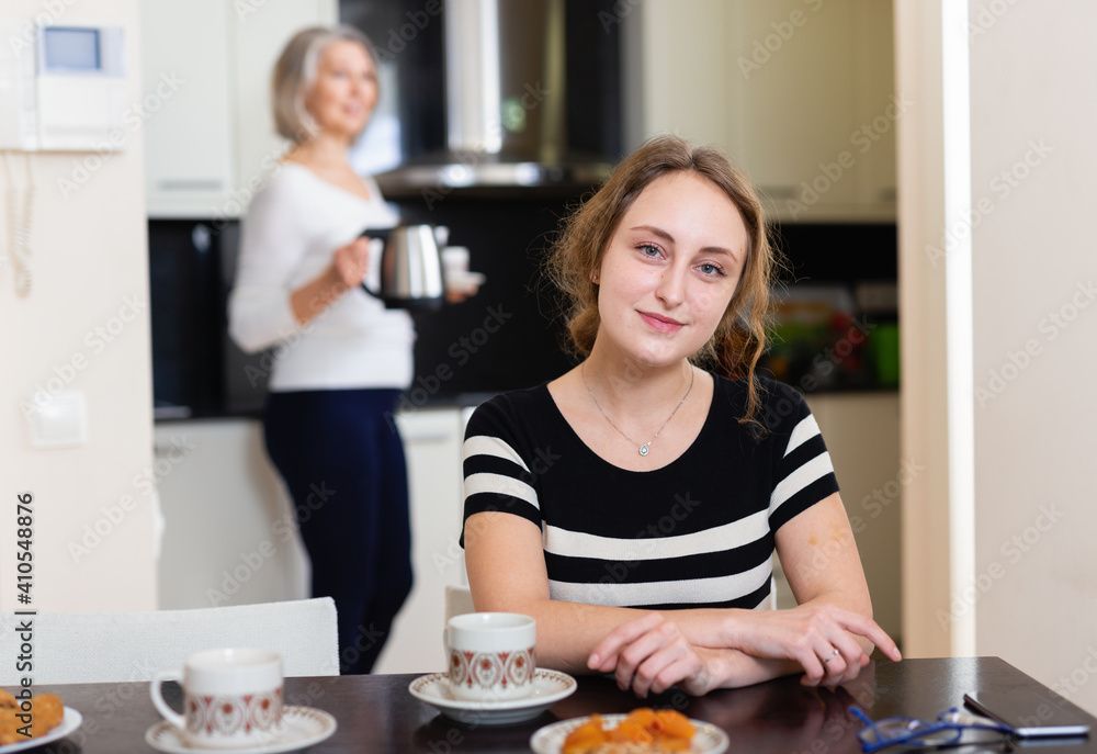 Smiling young woman enjoying tea at kitchen table, elderly mother at background