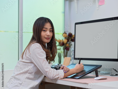 Female office worker smiling to camera while working with drawing tablet and computer