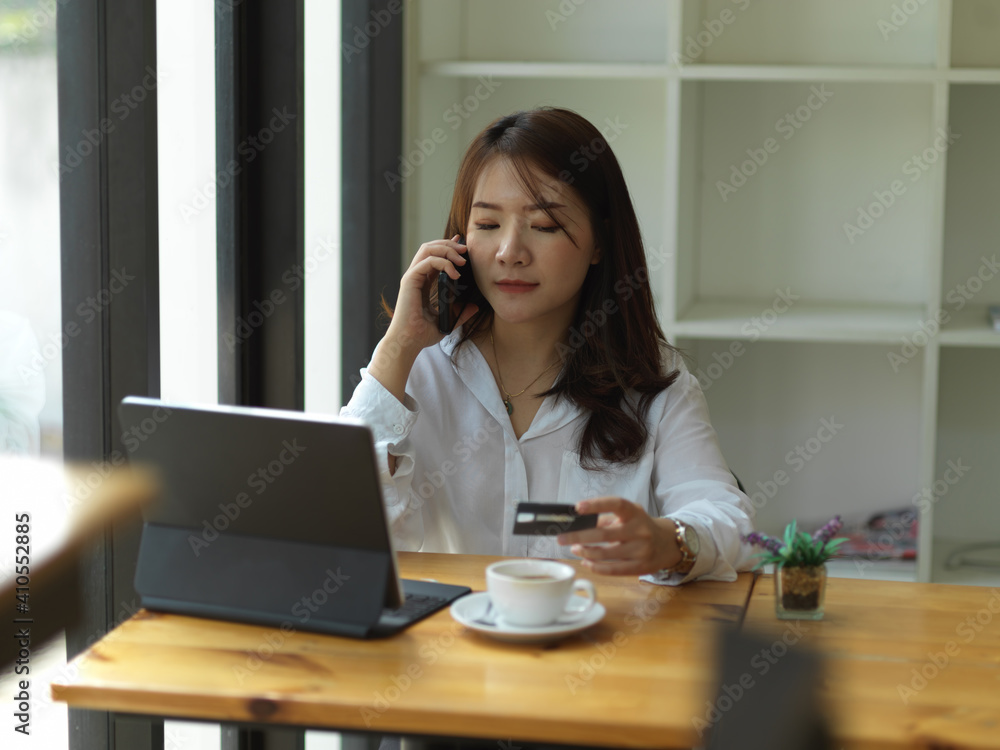 Female freelancer talking on the phone and holding credit card while working in cafe Female freelancer talking on the phone and holding credit card while working in cafe