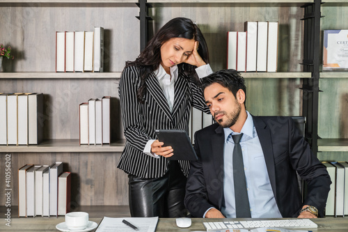 A young businesswoman in a suit showing a tablet screen to a businessman sitting on his desk in an office looking disappointed, unhappy, and worry. photo