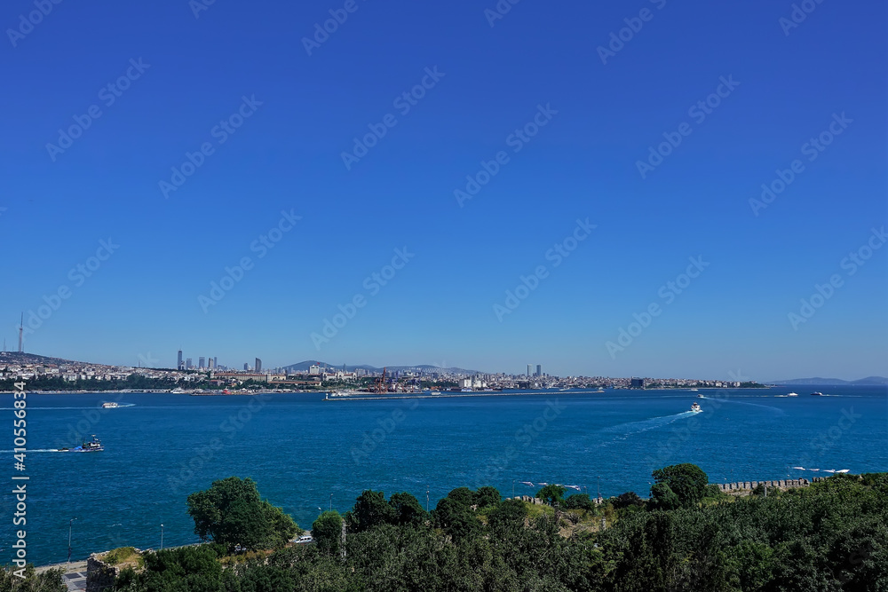 Ships sail on the blue surface of the Bosphorus. Residential buildings, Istanbul skyscrapers, silhouettes of mountains are visible in the distance. Green trees in the foreground.