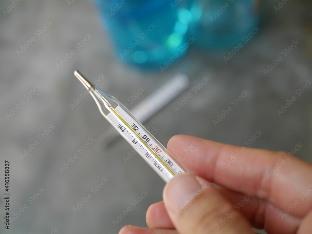 Hand held a close-up of a glass thermometer. Behind there is a picture of hand washing alcohol during the Covid-19 outbreak.