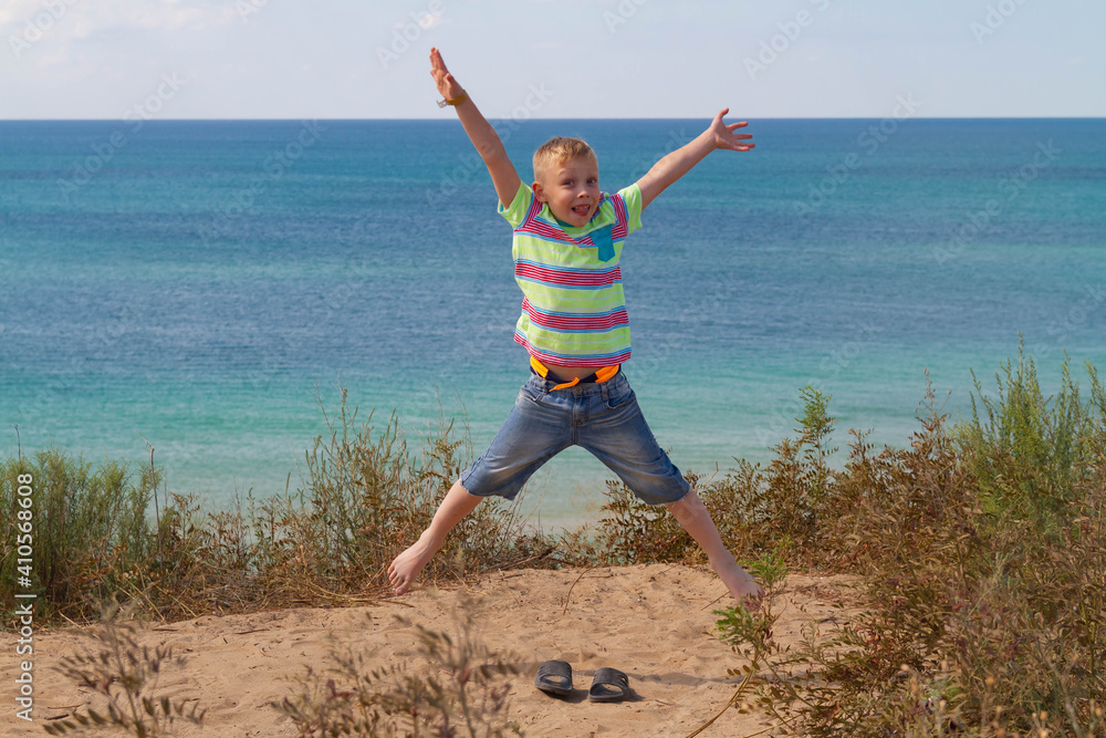 A blond European boy jumps high on a hill by the sea and smiles.