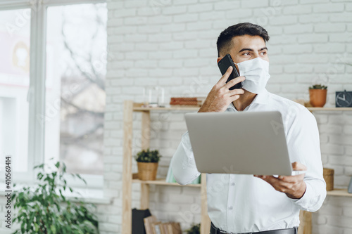 Handsome man businessman in medical mask using his laptop while talking on his cellphone