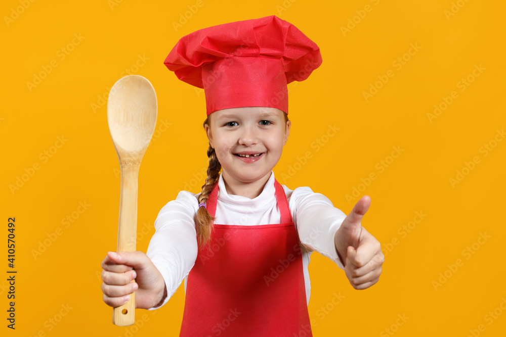 Cute child chef on a yellow background. A little girl holds a wooden spoon in her hands and shows her thumb up. Cute child chef on a yellow background. A little girl holds a wooden spoon in her hands and shows her thumb up.