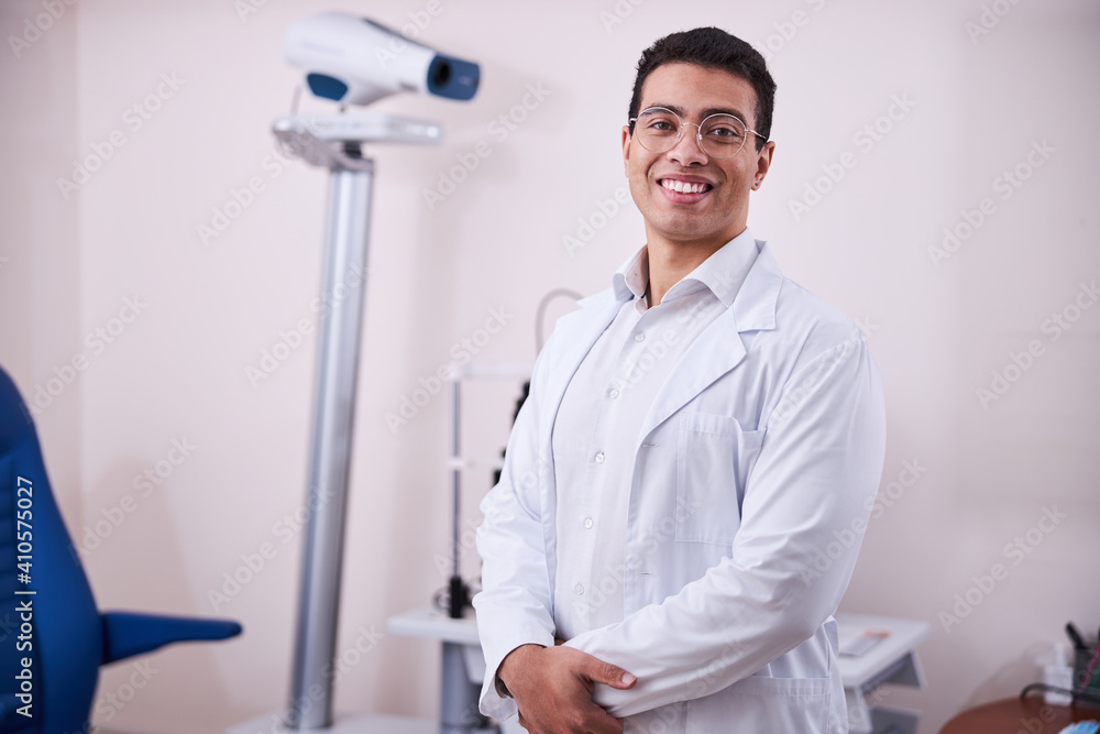 Cheerful bespectacled eye doctor standing in his office