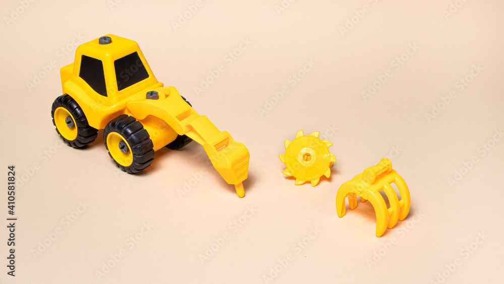 Obraz premium Children's plastic toy on a beige background - a yellow tractor with different nozzles drill, saw on wood, a wood loader, a bucket excavator. Construction equipment for children.