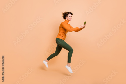 Full length photo of funny cute young lady wear orange turtleneck jumping typing modern device isolated beige color background