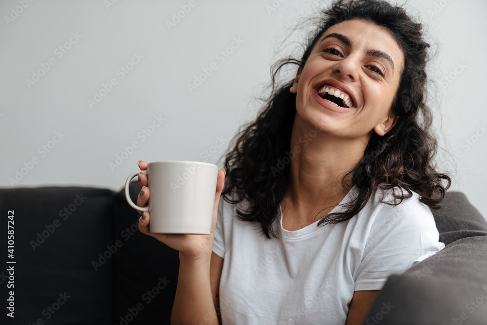 Happy brunette woman laughing and drinking coffee while sitting on sofa Happy brunette woman laughing and drinking coffee while sitting on sofa