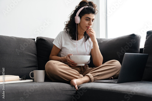 Focused brunette woman in headphones using laptop while eating cereal