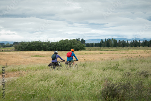 Two cyclists riding the Otago Central Rail Trail among the field of long grass blowing in wind, South Island, New Zealand
