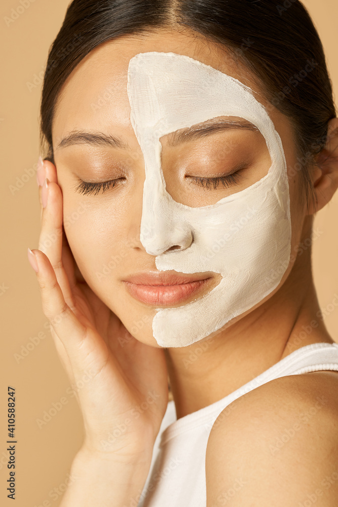 Relaxed young woman with facial mask applied on half of her face receiving spa treatments, posing for camera with eyes closed isolated over beige background Relaxed young woman with facial mask applied on half of her face receiving spa treatments, posing for camera with eyes closed isolated over beige background