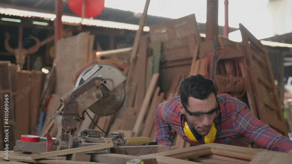 Young carpenter wearing safety goggle using sandpaper to polish workpiece to smooth in furniture manufacturing and blow the dust off. Craftsman standing working in workplace with tool and equipment. Young carpenter wearing safety goggle using sandpaper to polish workpiece to smooth in furniture manufacturing and blow the dust off. Craftsman standing working in workplace with tool and equipment.