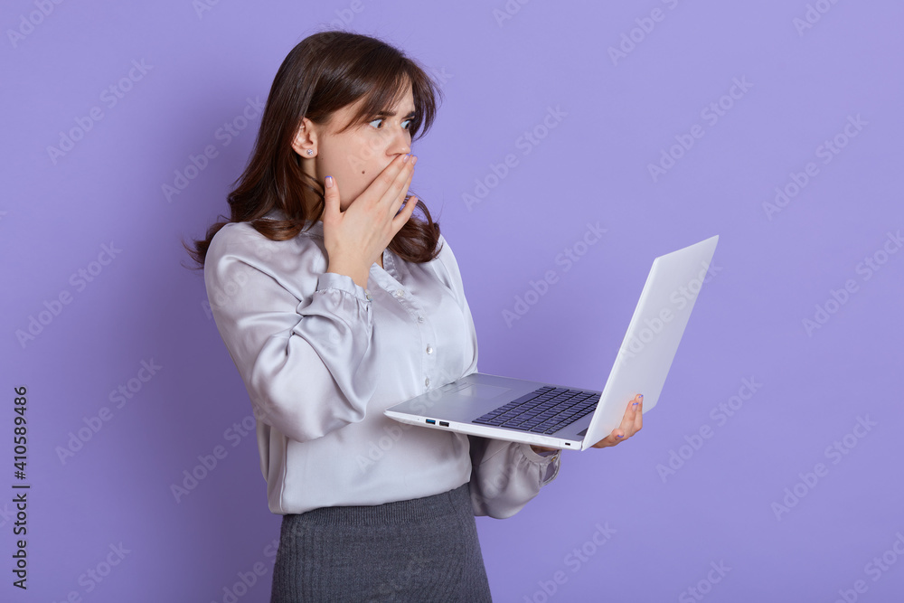 Attractive shocked businesswoman with laptop in hands posing isolated over lilac background, wearing elegant attire, has unexpected trouble with portable computer or her work, covers mouth with palm. Attractive shocked businesswoman with laptop in hands posing isolated over lilac background, wearing elegant attire, has unexpected trouble with portable computer or her work, covers mouth with palm.