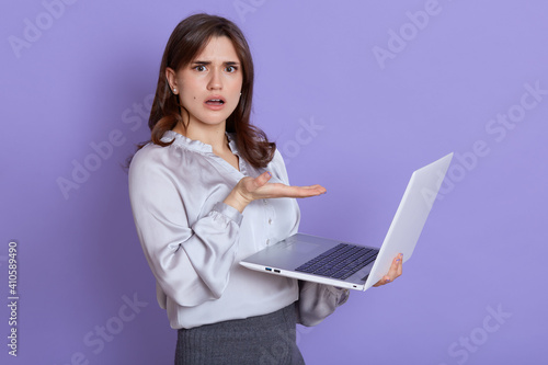 Young European woman frustrated by computer operational problems, looking at camera with puzzled and astonished expression, has unexpected troubles, pointing at laptop screen with palm.