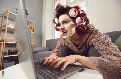 Fotografie, Obraz Mad woman in face mask and hair rollers sitting looking at laptop computer screen, writing insulting hater comment, spreading fake rumour