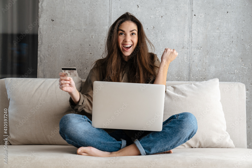 Delighted girl winking and holding credit card while using laptop at home Delighted girl winking and holding credit card while using laptop at home