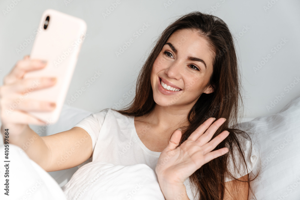 Happy nice girl waving hand while using mobile phone in bed at home Happy nice girl waving hand while using mobile phone in bed at home