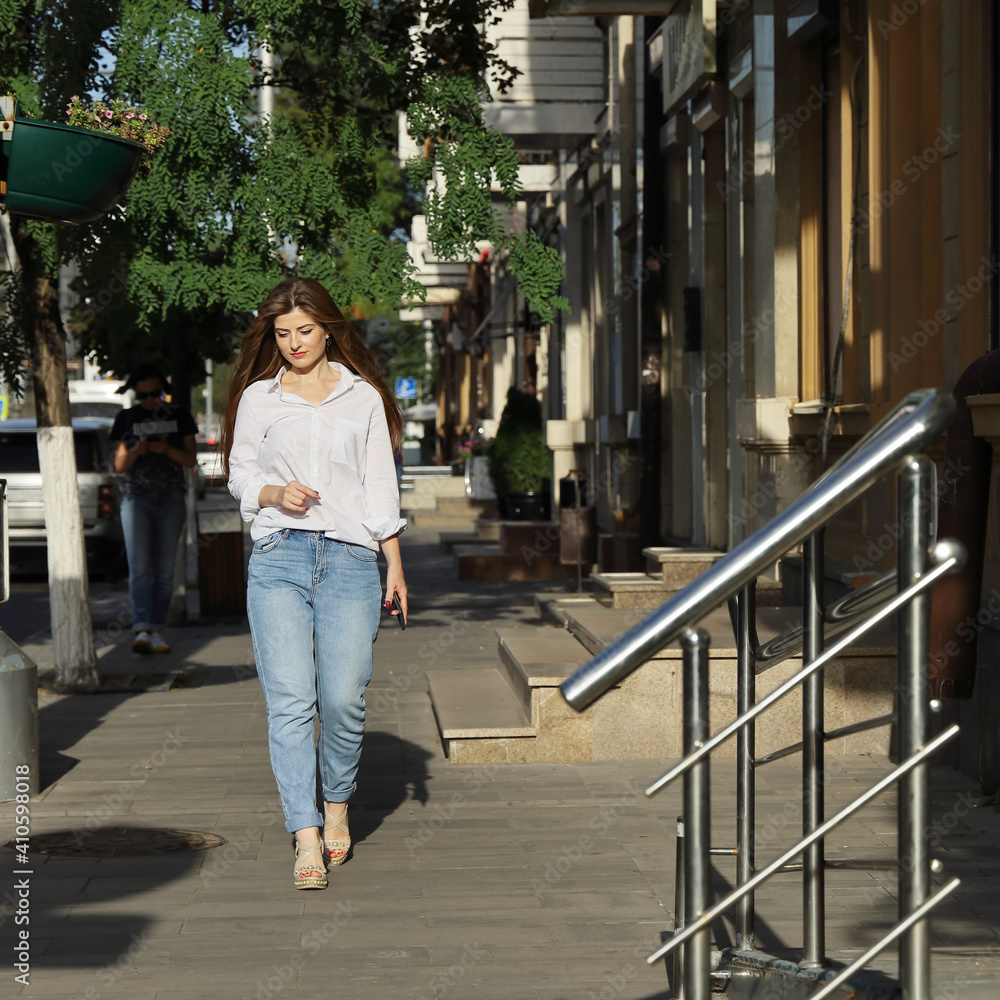 Young beautiful woman in white blouse and jeans walks slowly down the street on summer sunny day