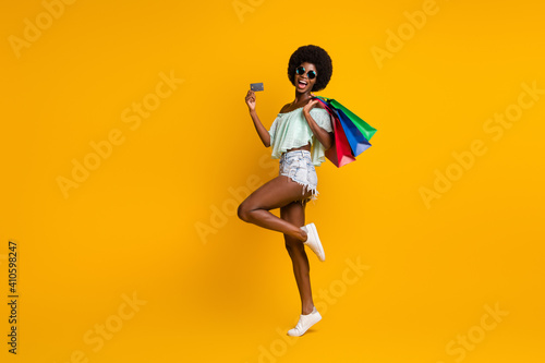 Full length body size photo of black skinned girl keeping bags showing plastic debit card isolated on vibrant yellow color background copyspace