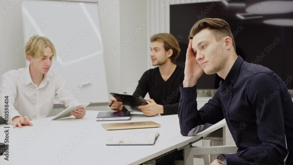 Side view of bored young Caucasian man sitting with coworkers in conference room sighing and touching face. Portrait of exhausted overworked employee in office indoors. Side view of bored young Caucasian man sitting with coworkers in conference room sighing and touching face. Portrait of exhausted overworked employee in office indoors.