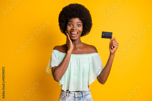Photo portrait of shocked woman touching face cheek holding debit card in one hand isolated on vivid yellow colored background