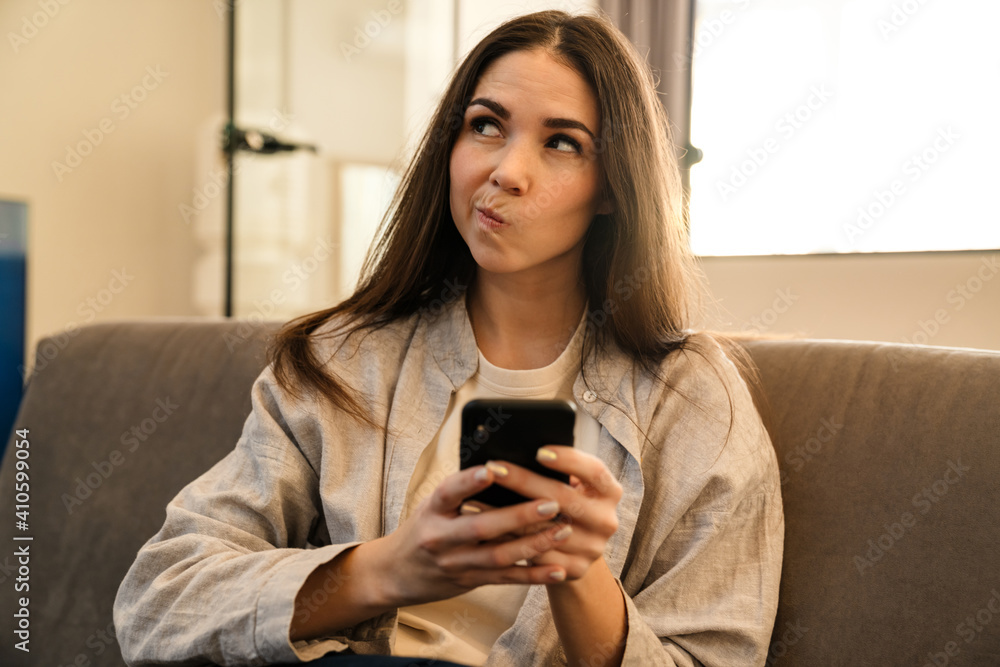 Perplexed young woman using smartphone while sitting on sofa indoors Perplexed young woman using smartphone while sitting on sofa indoors