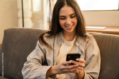 Happy young woman smiling and using smartphone while sitting on sofa