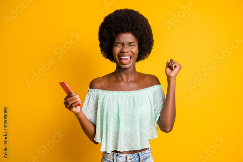 Photo portrait of screaming girl celebrating with fist up holding phone in one hand isolated on vivid yellow colored background