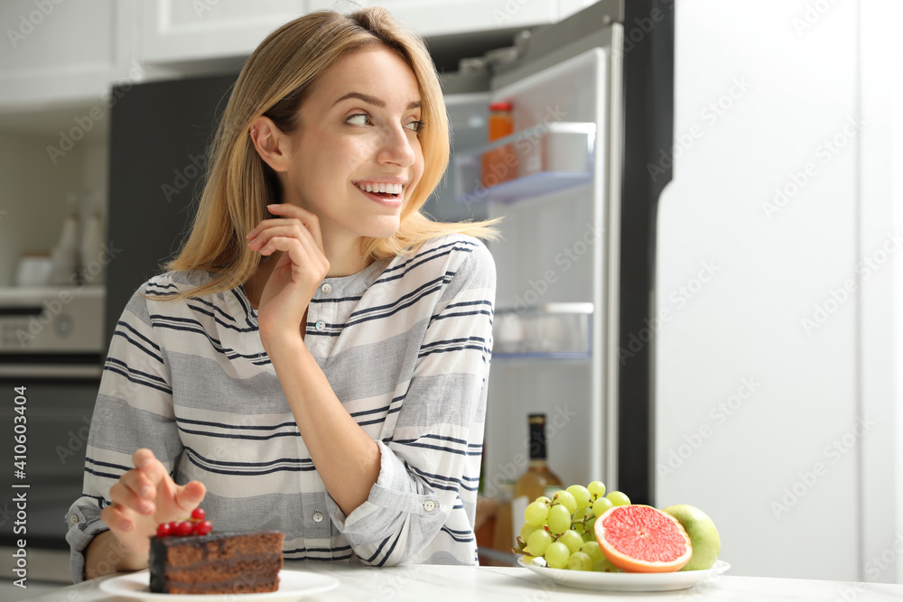 Woman choosing between cake and healthy fruits at table in kitchen