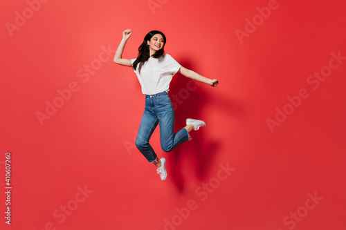 Mischievous girl in white T-shirt and jeans jumping on red background