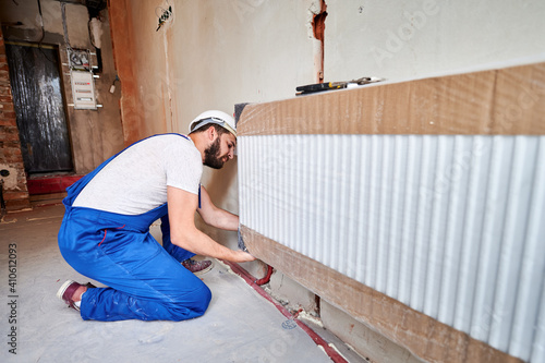 Male plumber in work overalls installing heating radiator in empty room. Bearded young man in safety helmet installing heating system in apartment. Concept of radiator installation, plumbing works. © anatoliy_gleb