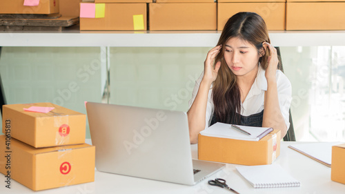 Start a small business in a cardboard box of a working Asian woman looking at a laptop screen dazed by customer orders and deliveries for customers selling things online.