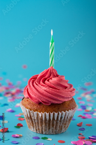 Pink birthday cupcake with green candle and colorful confetti on blue background