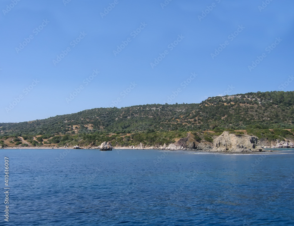 View of the coast of Cyprus from the Mediterranean Sea. A rocky coastline stretches along the calm blue water. Rocks protrude into the sea. Green vegetation on the mountain. Clear azure sky.