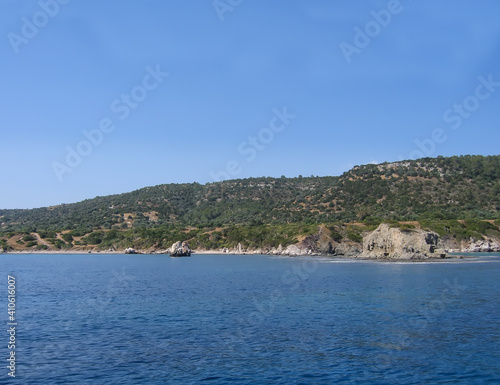 View of the coast of Cyprus from the Mediterranean Sea. A rocky coastline stretches along the calm blue water. Rocks protrude into the sea. Green vegetation on the mountain. Clear azure sky.