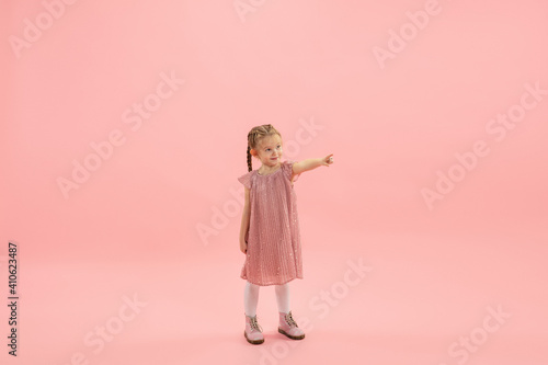 Pointing. Childhood and dream about big and famous future. Pretty longhair girl on coral pink studio background. Childhood, dreams, imagination, education, facial expression, emotions concept.