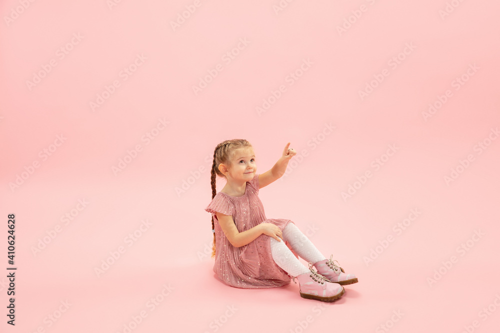 Sitting, pointing. Childhood and dream about big and famous future. Pretty longhair girl on coral pink studio background. Childhood, dreams, imagination, education, facial expression, emotions concept Sitting, pointing. Childhood and dream about big and famous future. Pretty longhair girl on coral pink studio background. Childhood, dreams, imagination, education, facial expression, emotions concept