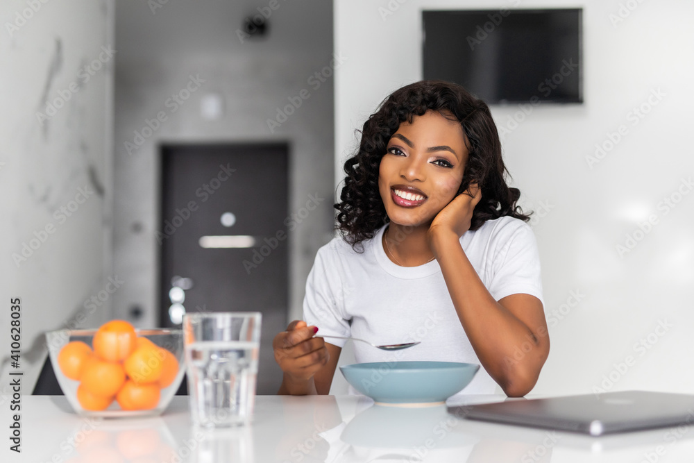 Smiling young african woman having breakfast while sitting at the kitchen with laptop Smiling young african woman having breakfast while sitting at the kitchen with laptop
