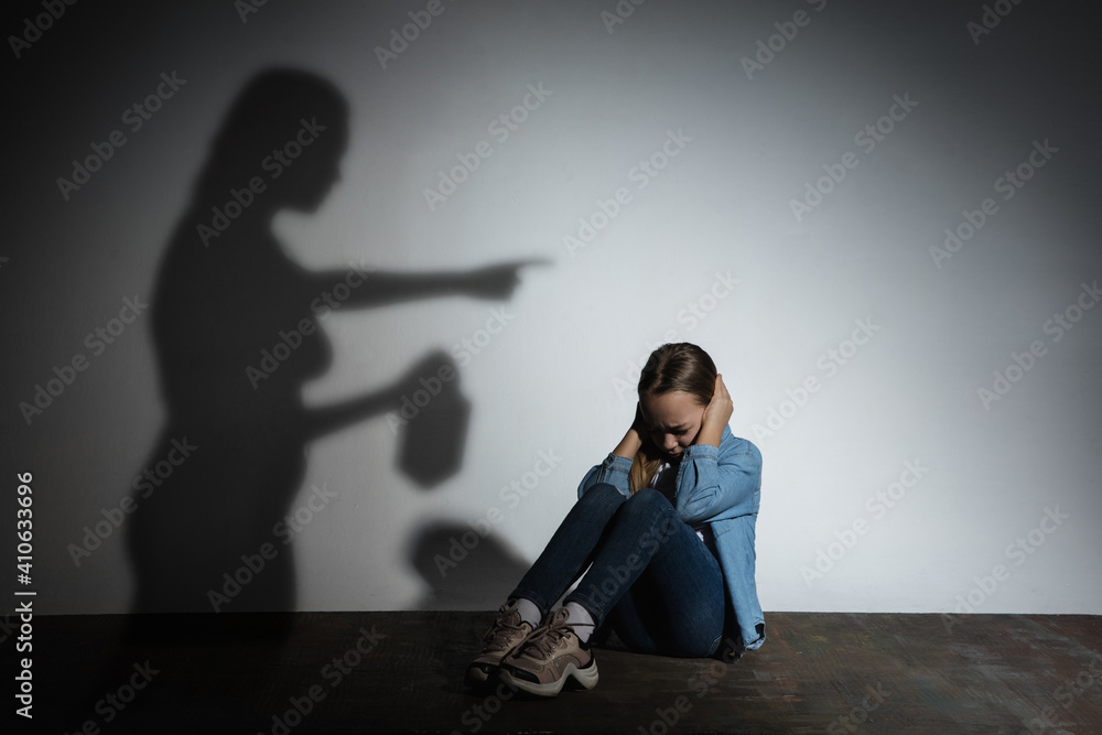 Domestic physical violence, abusing. Scared little caucasian girl, victim sitting close to white wall with shadow of angry threatening mother with alcohol addiction. Awareness of social problem. Domestic physical violence, abusing. Scared little caucasian girl, victim sitting close to white wall with shadow of angry threatening mother with alcohol addiction. Awareness of social problem.