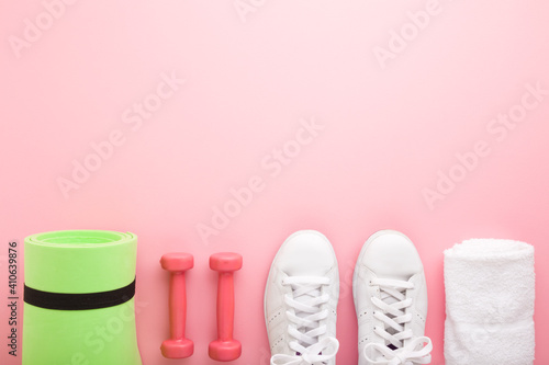 Female fitness training accessories. White sport shoes, dumbbells, green foam mat and towel on light pink table. Pastel color. Closeup. Empty place for motivational, inspirational text. Top view.