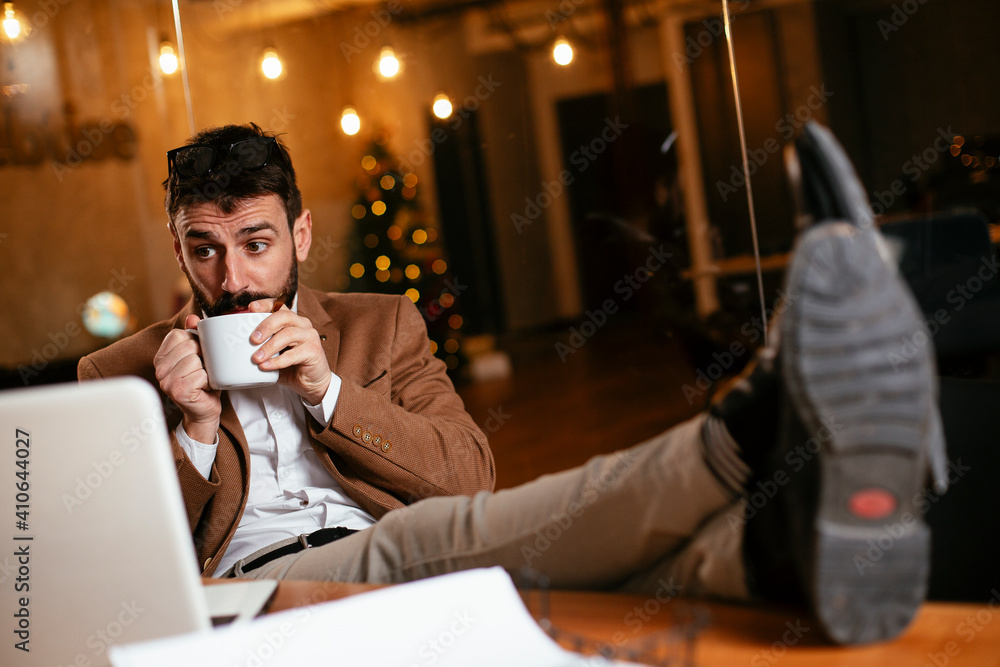Obraz premium Young businessman using laptop and drinking coffee in his office. Handsome man on coffee break