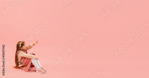 Pointing. Childhood and dream about big and famous future. Pretty longhair girl on coral pink studio background. Childhood, dreams, imagination, education, facial expression, emotions concept. Flyer