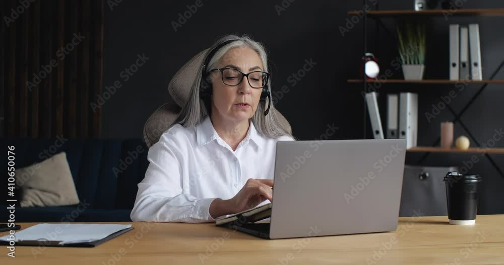 Old Mature Senior Woman Teacher, Lecturer, Coach is wearing Headphones with Microphone, holding a Seminar, Conference, Webinar. Progressive old Female Lecturer working Indoor on Quarantine. Education. Old Mature Senior Woman Teacher, Lecturer, Coach is wearing Headphones with Microphone, holding a Seminar, Conference, Webinar. Progressive old Female Lecturer working Indoor on Quarantine. Education.
