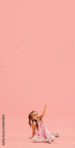 Pointing. Childhood and dream about big and famous future. Pretty longhair girl on coral pink studio background. Childhood, dreams, imagination, education, facial expression, emotions concept. Flyer