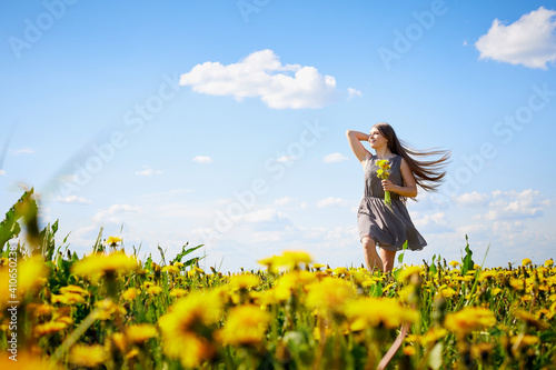 Beautiful young woman on a field with green grass and yellow dandelion flowers in a sunny day. Girl on nature with yellow flowers and blue sky.