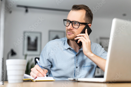 A busy young entrepreneur wearing glasses and smart casual shirt is talking on the mobile phone and using a laptop. Remote work concept