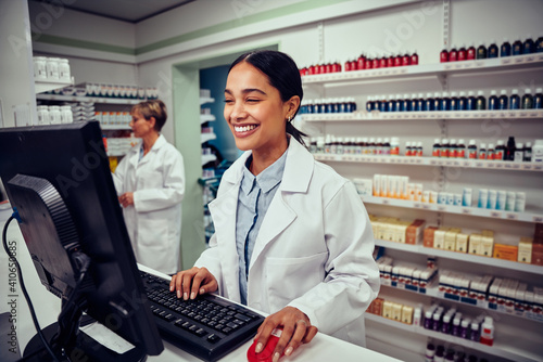 Cheerful young female pharmacist wearing labcoat working on computer in drugstore photo