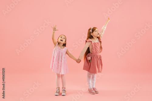 Pointing. Childhood and dream about big and famous future. Pretty little girls on coral pink studio background. Childhood, dreams, imagination, education, facial expression, emotions concept.